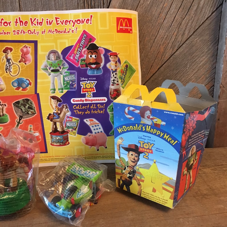Pode incluir: Uma caixa de Happy Meal do McDonald's com o Woody do filme Toy Story 2. A caixa &eacute; amarela e vermelha com uma imagem do Woody na frente. O texto "McDonald's Happy Meal" est&aacute; impresso em branco sobre um fundo vermelho. O texto "Disney Pixar Toy Story 2" est&aacute; impresso em branco sobre um fundo amarelo. A caixa est&aacute; sobre uma mesa de madeira.