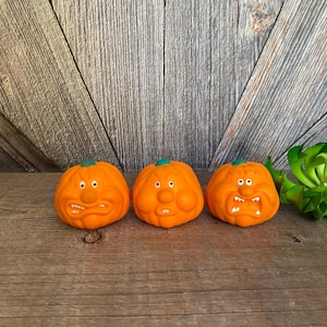 May include: Three orange, cartoon-like pumpkins with green stems and expressive faces. Each pumpkin has a unique mouth shape and white eyes. The pumpkins are arranged on a wooden surface, evoking a Halloween theme.
