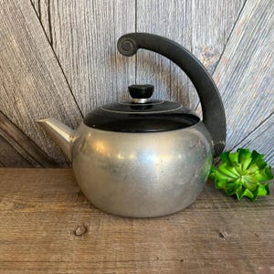 May include: A silver aluminum teapot with a black lid and handle. The teapot has a round body and a spout on the side.