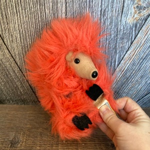 May include: A bright orange plush hedgehog toy with black feet and a brown nose. The toy is made of soft, fluffy fur.