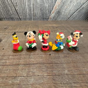 May include: A collection of five Christmas-themed Disney character figurines. Pluto is in a red and green stocking. Mickey Mouse, Minnie Mouse, and Donald Duck are dressed in festive attire. Each figurine is on a small, round base.