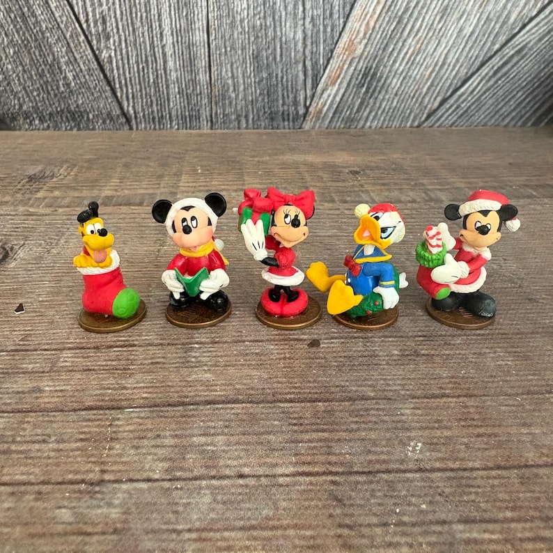 May include: A collection of five Christmas-themed Disney character figurines. Pluto is in a red and green stocking. Mickey Mouse, Minnie Mouse, and Donald Duck are dressed in festive attire. Each figurine is on a small, round base.