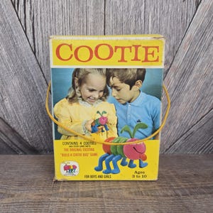 May include: Vintage "Cootie" game box with a yellow background and a yellow handle. The box features a picture of two children and a colorful Cootie bug. Text on the box reads "Cootie" and "Build a Cootie Bug Game". Ages 3 to 10.