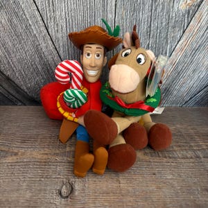 May include: A plush Woody doll with a brown cowboy hat, red shirt, and blue pants, holding a candy cane. Beside him is a plush Bullseye horse wearing a green wreath. The toys are set against a wooden background.