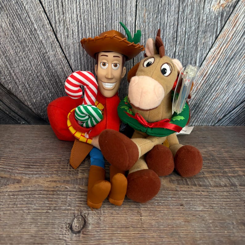 May include: A plush Woody doll with a brown cowboy hat, red shirt, and blue pants, holding a candy cane. Beside him is a plush Bullseye horse wearing a green wreath. The toys are set against a wooden background.