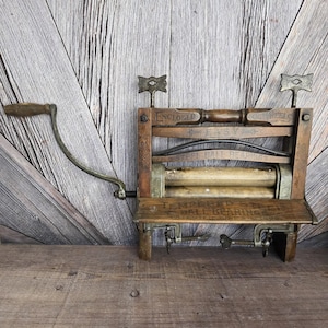 May include: Antique wooden and metal clothes wringer with a hand crank. The wringer has a wooden frame with metal rollers and clamps. The text "ENCLOSED", "EASY", and "STEEL BALL BEARINGS" are visible.