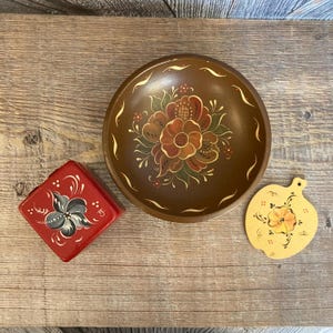 May include: Three hand-painted wooden bowls with floral designs. The bowl on the left is red with a black flower, the bowl in the center is brown with a red and yellow flower, and the bowl on the right is yellow with an orange flower.