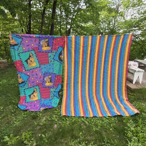 May include: Two colorful fabric panels hanging outdoors. One features a patchwork design with a Native American woman in a yellow dress. The other has vertical stripes in blue, yellow, orange, and purple. The fabrics are draped over a clothesline.
