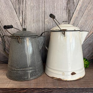 May include: Two vintage enamelware coffee pots. One is speckled gray, the other is white with black trim. Both have lids, spouts, and wire handles with black wooden grips. They sit on a wooden shelf against a wood-paneled background.