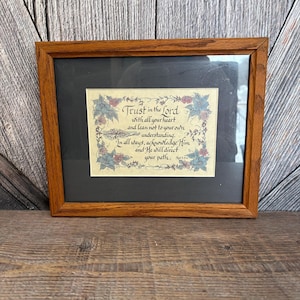 May include: Framed artwork with a wooden frame. The artwork features the text "Trust in the Lord" with floral and leaf accents in blue, pink, and green. The frame is a medium brown color.