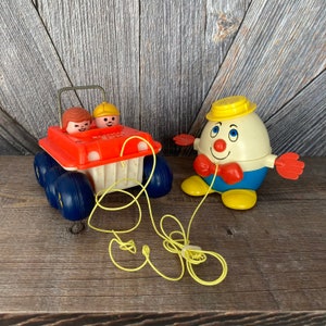May include: Two vintage Fisher-Price toys. An orange and white toy car with blue wheels and two toy figures inside. A yellow pull string is attached. Next to it is a Humpty Dumpty toy with a yellow hat and red bow tie.