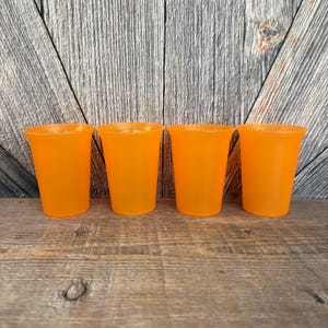 May include: Four orange plastic cups are arranged in a row. The cups have a slightly flared rim and a smooth, matte finish. The cups are set against a weathered wood background.