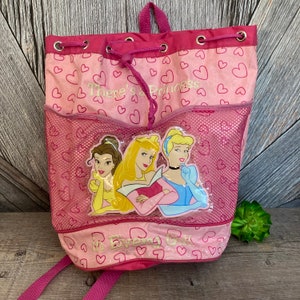 May include: Pink drawstring backpack with a mesh pocket featuring a graphic of three Disney princesses: Belle, Aurora, and Cinderella. The backpack has a pink and white heart pattern and the text "There's a Princess in Every Girl".