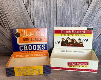 May include: Two vintage cigar boxes. The box on the left is blue, yellow, and orange with the text "Wolf Bros. Rum Soaked Crooks Dipped in Wine Wolf Bros. Crooks". The box on the right is white, red, and yellow with the text "Dutch Masters Perfectos 154 Dutch Masters Perfectos The Fine Cigar The Fine Cigar".