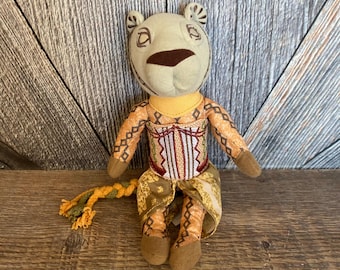 May include: A plush lion toy wearing a yellow and brown patterned dress with a red and gold embroidered bodice. The lion has a braided tail and a light brown face.