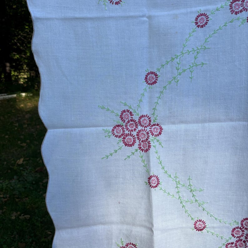 May include: White linen tablecloth with a scalloped edge, featuring embroidered pink flowers and green stems. The floral design is arranged in clusters and along curved lines, creating a delicate and vintage aesthetic.