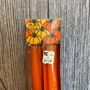 May include: A package of orange candles with a decorative top featuring pumpkins and the words "Pumpkin Time." The candles are in a clear plastic container with a price tag.