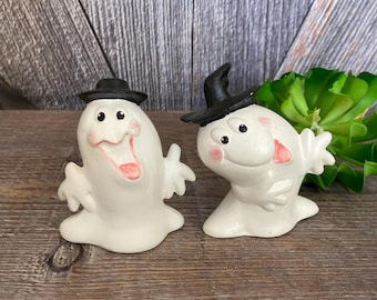 May include: Two white ceramic ghost figurines with black hats. The ghosts have pink cheeks and are smiling. One ghost is sticking its tongue out.