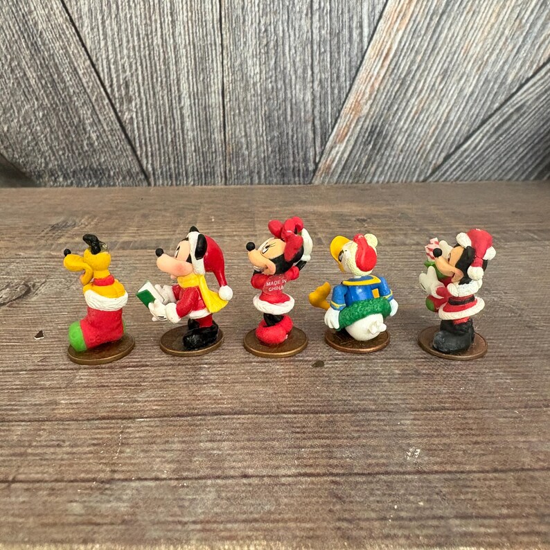 May include: A collection of five miniature Disney character figurines in festive Christmas attire. Pluto is in a stocking, Mickey Mouse is holding a book, Minnie Mouse is wearing a dress, Donald Duck is in a sweater, and Mickey and Minnie are together.