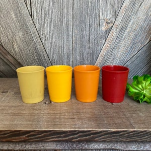 May include: Set of four plastic cups in shades of yellow, orange, and red. The cups are arranged in a row from left to right, with the yellow cup on the left and the red cup on the right.