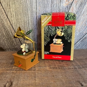 May include: A vintage-style, wooden, music box Christmas ornament with a gold-colored gramophone and two mice dancing on a revolving record. The ornament plays "Jingle Bells" and has a light that illuminates the gramophone. The box reads "Magic Motion! Music!" and "Song and Dance".