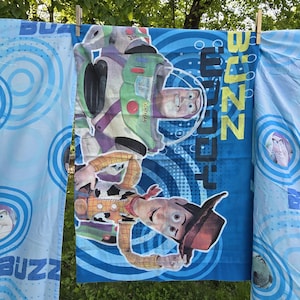 May include: Blue and white Toy Story themed bedsheets hanging on a clothesline. The sheets feature images of Buzz Lightyear and Woody, with the word "BUZZ" and "WOODY" in large letters. The background has a blue swirl design.