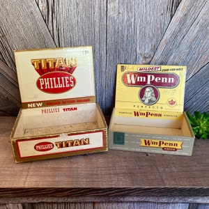 May include: Two vintage cigar boxes. The first box is gold and red with the text "TITAN PHILLIES" and "NEW BIGGER BETTER MILDER PHILLIES TITAN". The second box is yellow and red with the text "MILDEST CIGAR EVER MADE WmPenn" and "PERFECTO WmPenn PERFECTO".