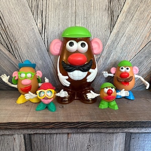 May include: A collection of Mr. Potato Head toys in various sizes and outfits. The toys have brown, tan, and orange bodies with accessories like hats, mustaches, and glasses. The toys are arranged on a wooden shelf against a wood-paneled background.