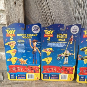 May include: Three Toy Story 2 action figure packages. The packages feature Woody and Buzz Lightyear, with the text "Silver Edition." The packages are blue, yellow, and red, with images of the characters and the movie logo.