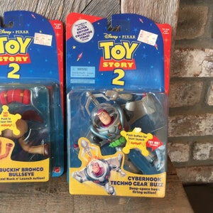 May include: Two Toy Story 2 action figures in original packaging. One features Buckin' Bronco Bullseye, and the other is Cyberhook Techno Gear Buzz. The packaging is blue and red with the Disney Pixar logo.