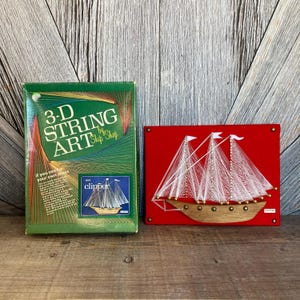 May include: A green box with the text "3-D String Art by Ship Shop" and a picture of a sailboat on the front. The box is next to a red felt board with a white string art sailboat on it. The sailboat is labeled "Clipper".