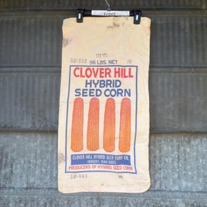 May include: Vintage Clover Hill Hybrid Seed Corn bag. The bag is beige with blue and red text and illustrations of four ears of corn. The bag reads "56 LBS. NET" and "Producers of Hybrid Seed Corn."