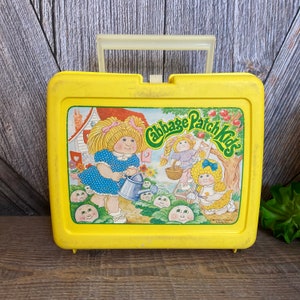 May include: Yellow plastic lunchbox with a Cabbage Patch Kids design. The lunchbox features a picture of three Cabbage Patch Kids dolls in a garden setting. The text "Cabbage Patch Kids" is printed in a red, white, and blue font.