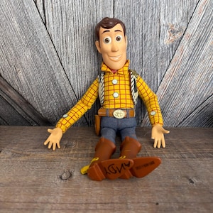May include: A Woody action figure from the Toy Story franchise. The figure is wearing a yellow plaid shirt, blue jeans, brown boots, and a brown belt with a gold buckle. The word "ANDY" is visible on the bottom of one boot.