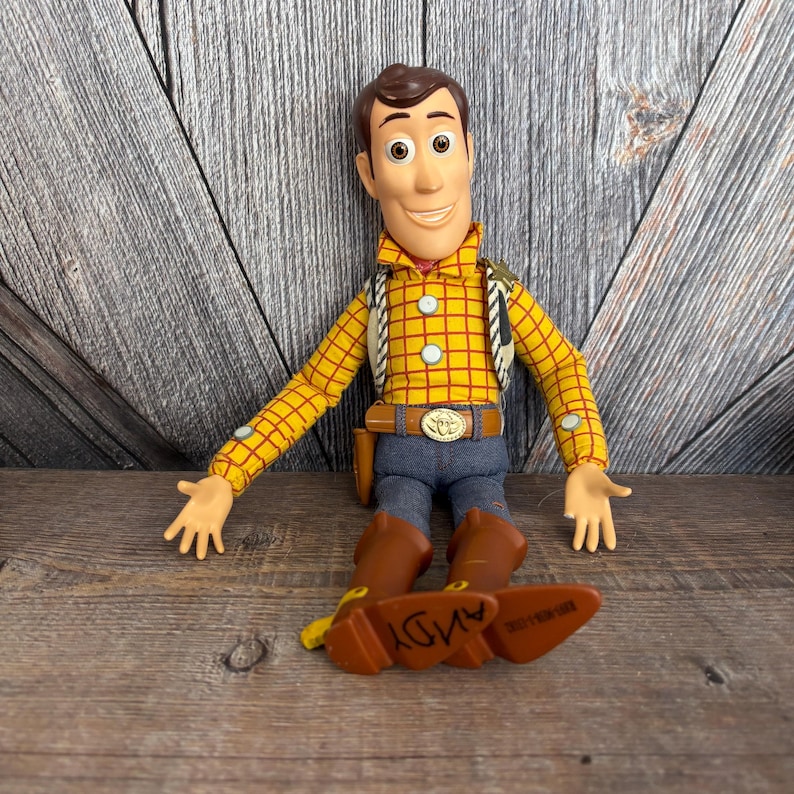 May include: A Woody action figure from the Toy Story franchise. The figure is wearing a yellow plaid shirt, blue jeans, brown boots, and a brown belt with a gold buckle. The word "ANDY" is visible on the bottom of one boot.