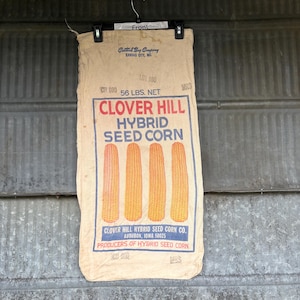 May include: A vintage Clover Hill Hybrid Seed Corn bag. The bag is beige with red and blue text, and features four stylized corn cobs. The bag reads "56 LBS. NET" and is from Audubon, Iowa.