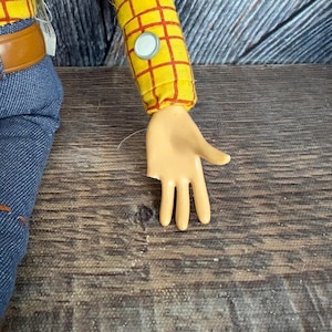 May include: A close-up of a Woody doll's arm and hand. The arm is covered in a yellow shirt with a red plaid pattern. The hand is tan and has three fingers. The doll is wearing blue jeans with a brown belt.