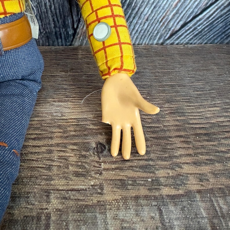 May include: A close-up of a Woody doll's arm and hand. The arm is covered in a yellow shirt with a red plaid pattern. The hand is tan and has three fingers. The doll is wearing blue jeans with a brown belt.