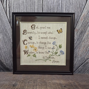 May include: Framed cross-stitch artwork with a black frame. The text reads "God, grant me Serenity, to accept what I cannot change, Courage, to change the things I can, & Wisdom, to know the difference. Amen." Includes floral and butterfly accents.