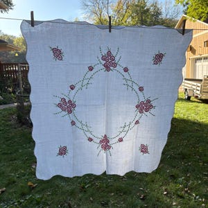 May include: White linen tablecloth with a scalloped edge, adorned with embroidered floral designs in shades of red and green. The embroidery forms a central wreath and scattered floral clusters.