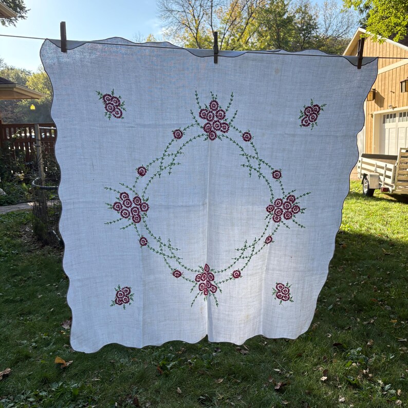 May include: White linen tablecloth with a scalloped edge, adorned with embroidered floral designs in shades of red and green. The embroidery forms a central wreath and scattered floral clusters.