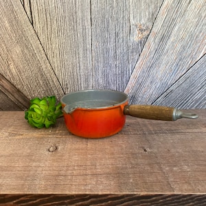 May include: Orange enamel cast iron saucepan with a wooden handle. The saucepan has a gray interior and a pouring spout.