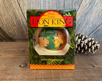 Puede incluir: Un adorno navideño en caja con Simba y Nala de El Rey León de Disney. El adorno es una bola de cristal transparente con una imagen colorida de los personajes en su interior. La caja es verde y presenta el logotipo de Disney y el título "Disney's The Lion King".