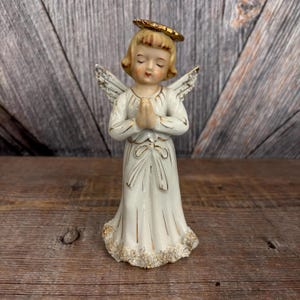 May include: A vintage ceramic angel figurine in a white gown with gold accents. The angel has closed eyes, folded hands, and a gold halo. The wings are white with gold detailing, and the base of the gown is trimmed with gold and white. The figurine is on a wooden surface.