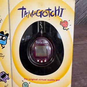 May include: A Tamagotchi virtual pet toy in its original packaging. The packaging is yellow and features the Tamagotchi logo and the text "the original virtual reality pet". The Tamagotchi toy is purple and has a small screen.