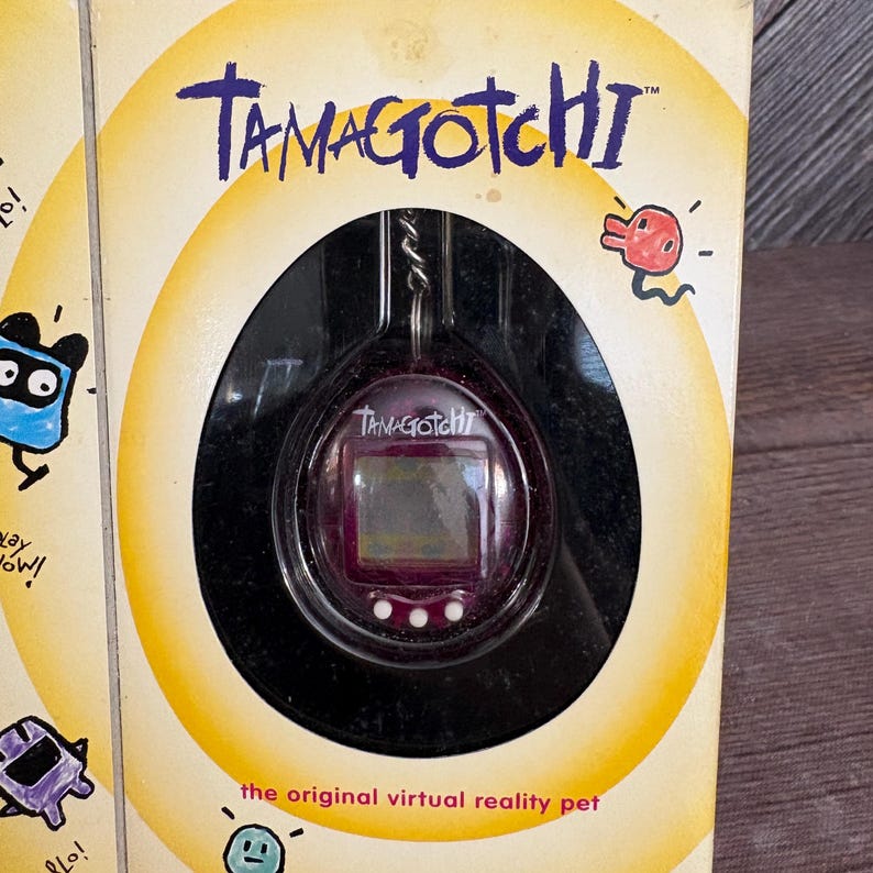 May include: A Tamagotchi virtual pet toy in its original packaging. The packaging is yellow and features the Tamagotchi logo and the text "the original virtual reality pet". The Tamagotchi toy is purple and has a small screen.
