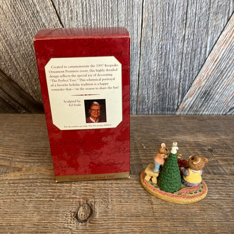 Pode incluir: Um ornamento de Natal feito &agrave; m&atilde;o com dois ursos a decorar uma pequena &aacute;rvore de Natal. O ornamento &eacute; datado de 1997 e foi esculpido por Ed Seale. "The Perfect Tree." Esta representa&ccedil;&atilde;o caprichosa de uma tradi&ccedil;&atilde;o natal&iacute;cia favorita &eacute; um alegre lembrete de que &eacute; a &eacute;poca para partilhar a divers&atilde;o!