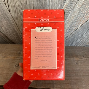 Puede incluir: Una caja roja y blanca con una caja de m&uacute;sica con un personaje de Disney en su interior. La caja presenta un logotipo de Disney y el texto "Schmid se enorgullece de presentarte esta caja de m&uacute;sica Walt Disney Character como parte de una colecci&oacute;n exclusiva de regalos hechos a mano, pintados a mano, que representan muchos a&ntilde;os de tradici&oacute;n Disney legendaria. Aqu&iacute; est&aacute;n todas las caras familiares con las que creciste, fielmente recreadas en cada detalle delicado. Y su encanto magn&eacute;tico cobra vida cuando tocan una melod&iacute;a favorita que se adapta perfectamente al estado de &aacute;nimo."