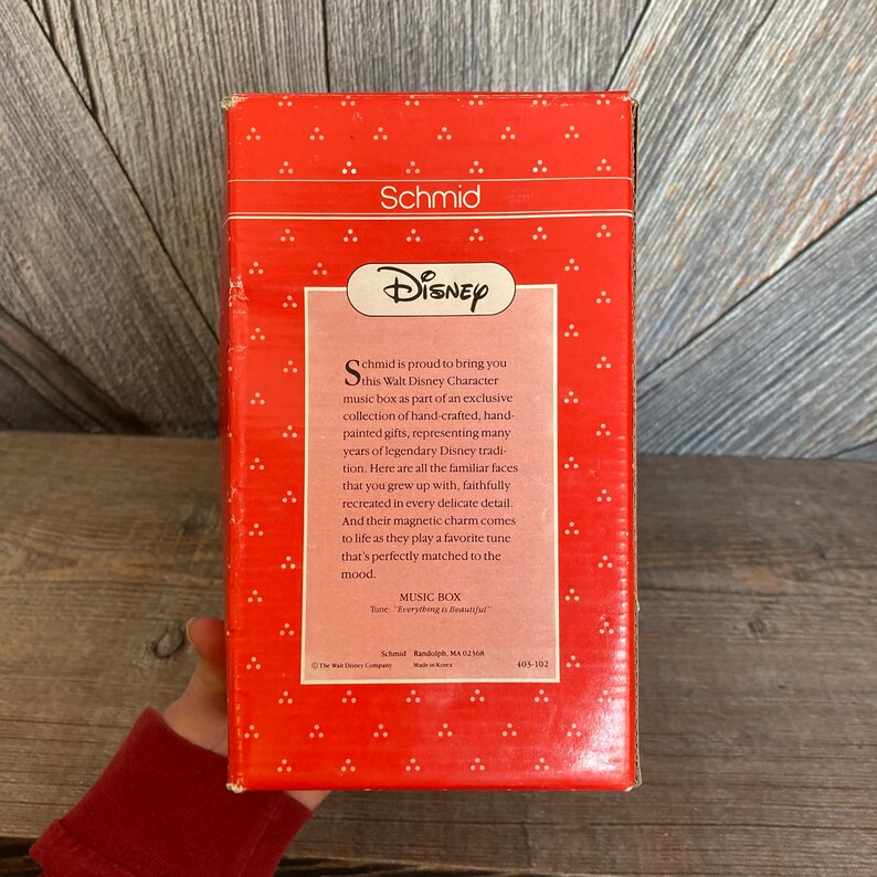 Puede incluir: Una caja roja y blanca con una caja de m&uacute;sica con un personaje de Disney en su interior. La caja presenta un logotipo de Disney y el texto "Schmid se enorgullece de presentarte esta caja de m&uacute;sica Walt Disney Character como parte de una colecci&oacute;n exclusiva de regalos hechos a mano, pintados a mano, que representan muchos a&ntilde;os de tradici&oacute;n Disney legendaria. Aqu&iacute; est&aacute;n todas las caras familiares con las que creciste, fielmente recreadas en cada detalle delicado. Y su encanto magn&eacute;tico cobra vida cuando tocan una melod&iacute;a favorita que se adapta perfectamente al estado de &aacute;nimo."