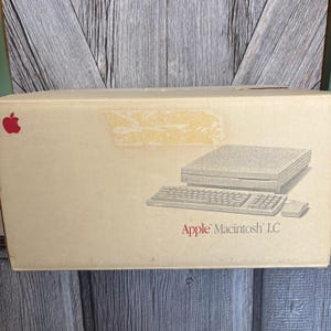 May include: A vintage Apple Macintosh LC computer box. The box is tan with a red Apple logo in the upper left corner. The box features a printed illustration of the computer, keyboard, and mouse. The text "Apple Macintosh LC" is printed in red.